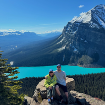 Two people sitting on a rock in front of a blue lake and mountains