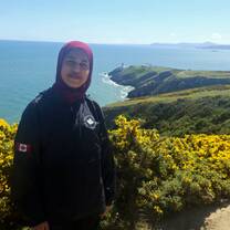 Lady in a hijab standing facing the camera behind her is a scenic view of the ocean and green hills