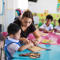A volunteer leans in to help a young child complete a wooden puzzle on a blue table in a vibrant preschool classroom.