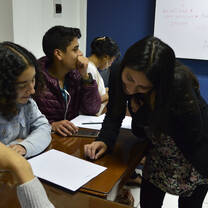 A woman leaning over a table with papers while seated learners watch.