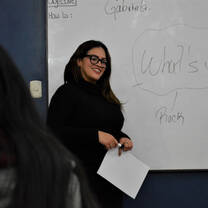 A smiling woman holding a paper and a marker in front of a whiteboard.