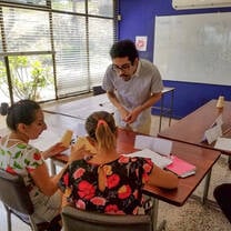 A man assisting two seated women during a cup-stacking classroom activity.
