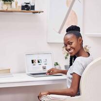 A smiling woman seated at a white desk with books and an open laptop.