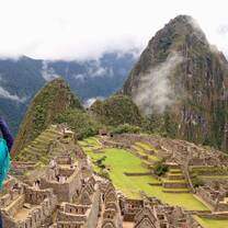 A traveller making a heart shape with raised hands in front of mountain ruins.