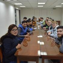 A group of people sitting at long wooden classroom tables.