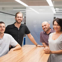 four people smiling and standing around a wooden table in front of a white board