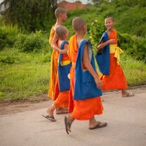 three monks wearing orange robes holding blue bags walking in Thailand